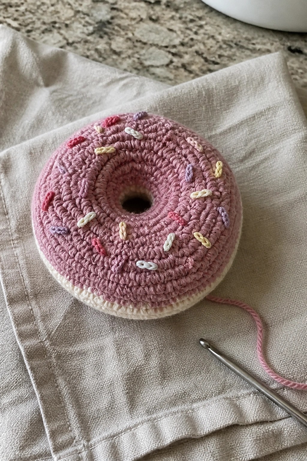 Pink crocheted donut with sprinkles on beige cloth beside crochet hook