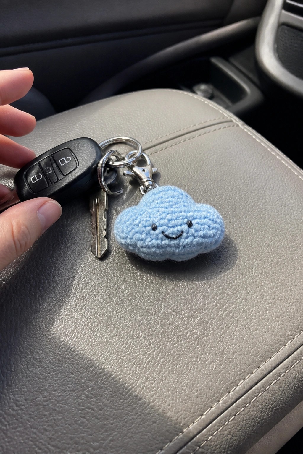 Hand holding car key fob and smiling blue crocheted cloud keychain on gray seat
