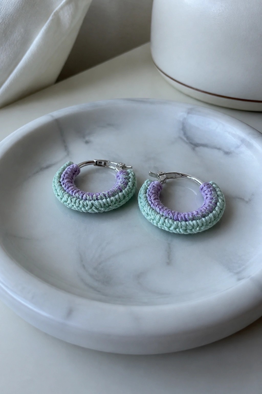 Two silver hoop earrings with mint green and purple crochet covers on a marble dish.