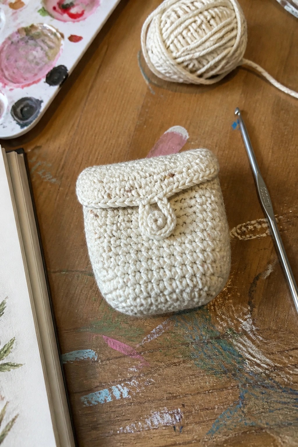 Cream crocheted pouch with button on paint-splattered wooden table beside yarn and hook