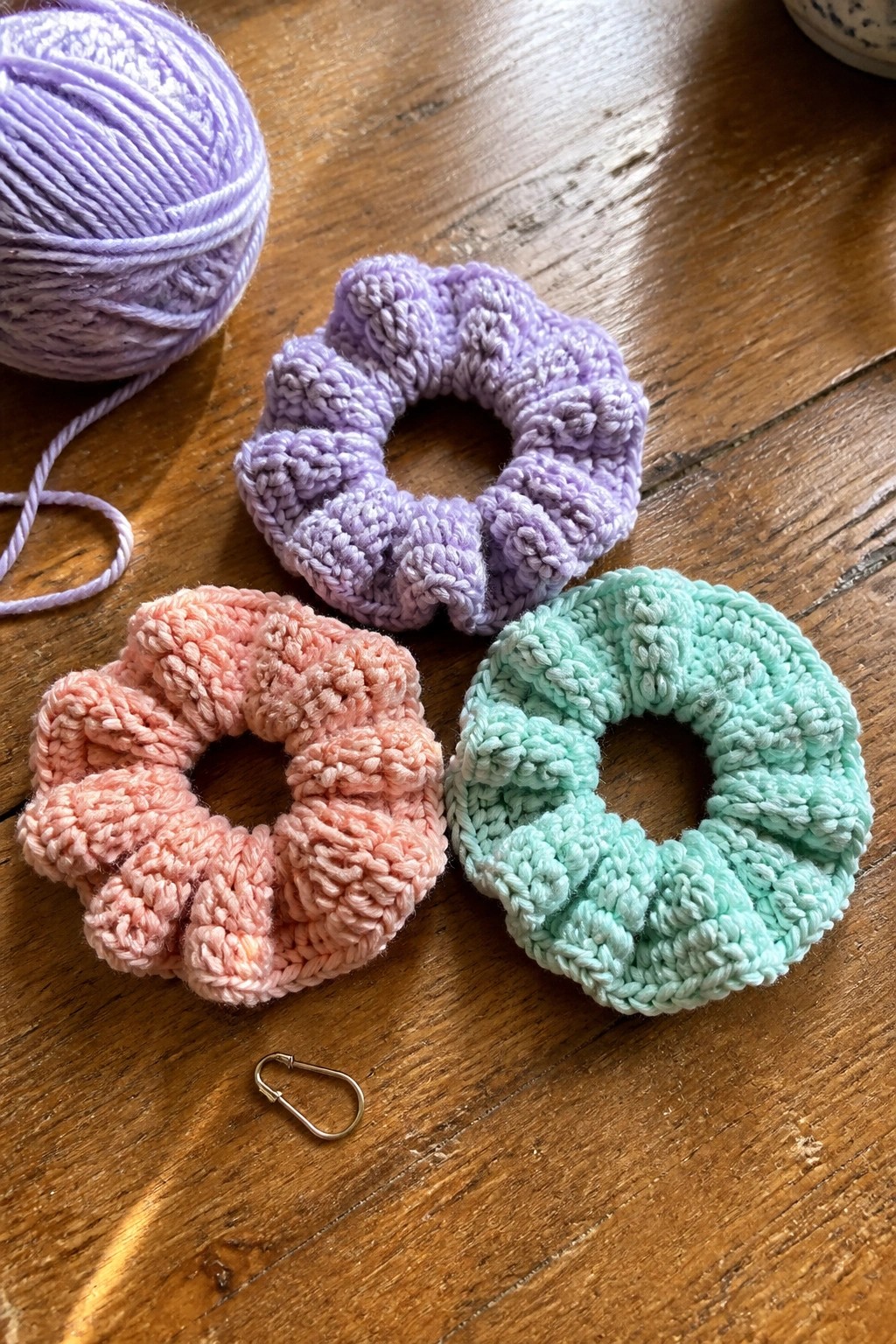 Three pastel crocheted scrunchies in purple, pink, and mint with yarn on wood.