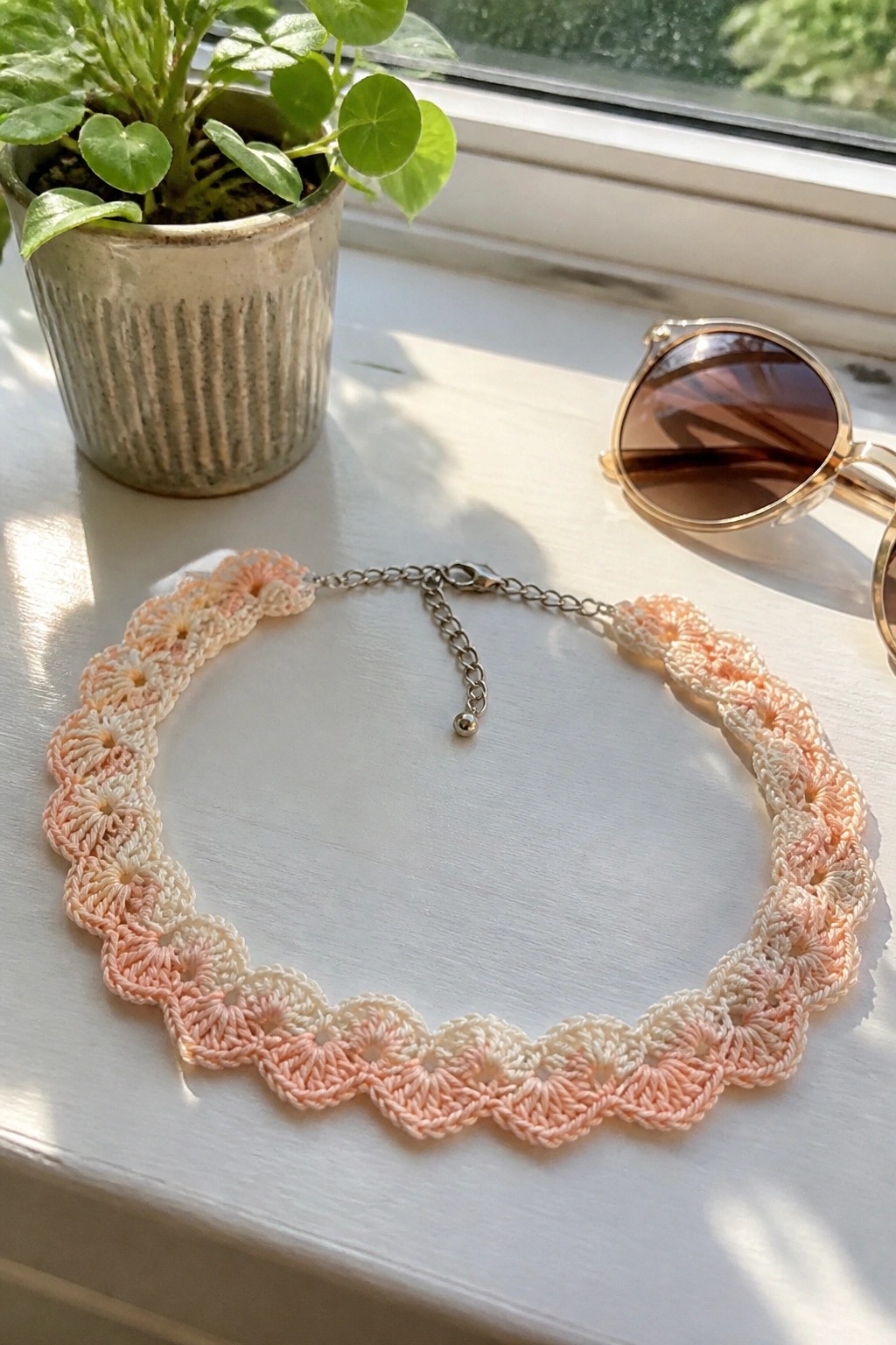 Crocheted peach gradient necklace on windowsill beside plant and sunglasses