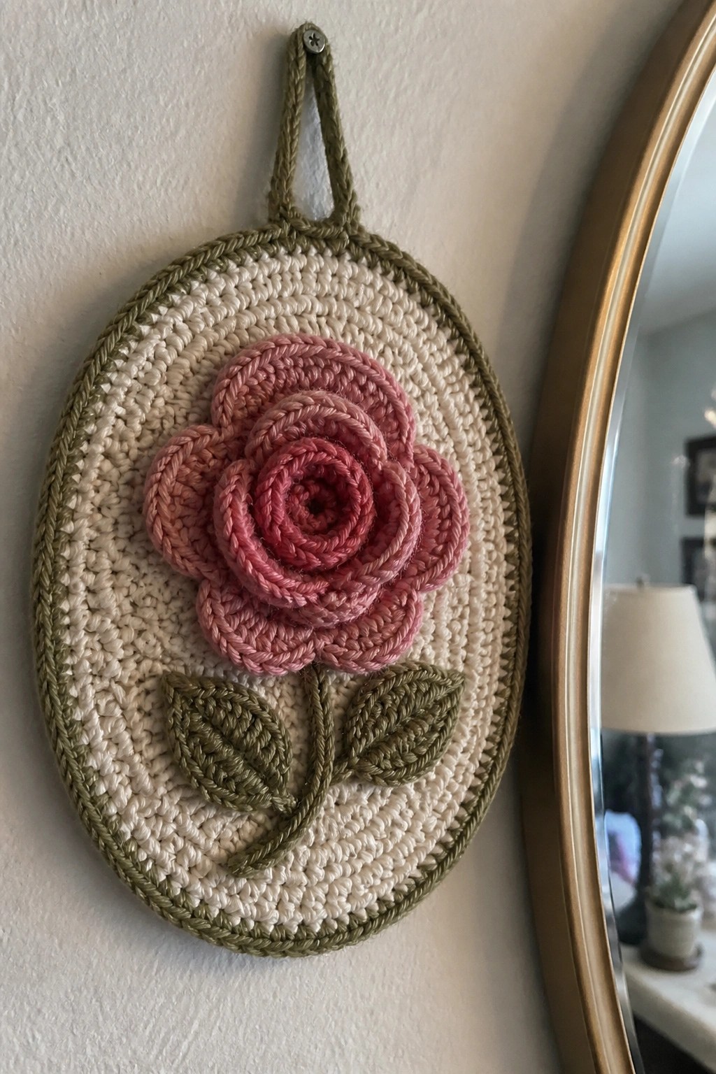 Oval crocheted wall hanging with pink rose, green leaves, and hanging loop.